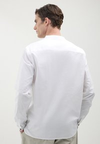 Weißes Langarmshirt aus leichtem Stoff, mit einem Knopfverschluss an den Manschetten und einem abgerundeten Saum. Schlichtes und sauberes Rückendesign.