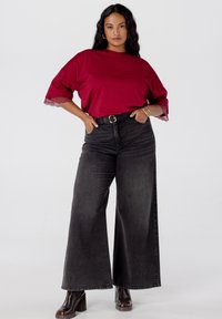 Vrouw met lang donker haar, gekleed in een rode top met kanten mouwen, hoge taille wijdvallende zwarte jeans, zwarte riem en zwarte hakken, met haar handen in de zakken.