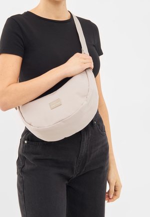 Osoba w czarnej koszulce i ciemnych dżinsach trzyma jasnobeżową torbę na ramię z markową metką na klatce piersiowej na białym tle.