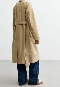 Persona con un abrigo trinchera beige con el cinturón atado en la espalda, pantalones vaqueros de pierna ancha azules y zapatillas blancas, de pie sobre un suelo gris contra una pared blanca.