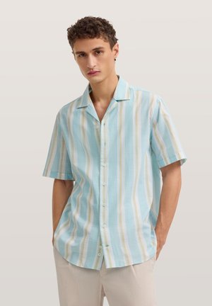 Junger Mann mit kurzärmeligem Hemd mit vertikalen blauen, weißen und beigen Streifen, steht mit den Händen in den Taschen.