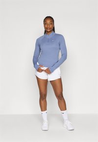 Langärmliges, hellblaues Sportoberteil mit halber Reißverschlus, kombiniert mit weißen Shorts. Verfügt über ein dezentes Nike-Logo auf der Brust.