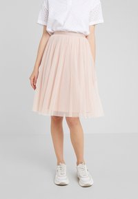 Mujer vistiendo una blusa blanca de manga corta perforada, una falda de tul rosa claro a la altura de la rodilla y zapatillas blancas, de pie frente a un fondo liso.