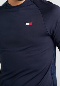 T-shirt de sport bleu marine à manches courtes, avec un petit logo rectangulaire comportant des bandes horizontales blanches et rouges sur la poitrine.