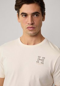 Krátky rukáv krémové tričko vyrobené z bavlny, obsahujúce malú vyšitú nášivku "H" s "HACKETT" pod ňou v kontrastnej farbe.