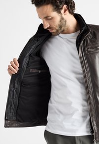 Bruine leren jas met ritssluiting, gevoerd met zwart quilted binnenwerk en binnenzak. De kraag is rechtopstaand en het ontwerp heeft een gladde textuur.