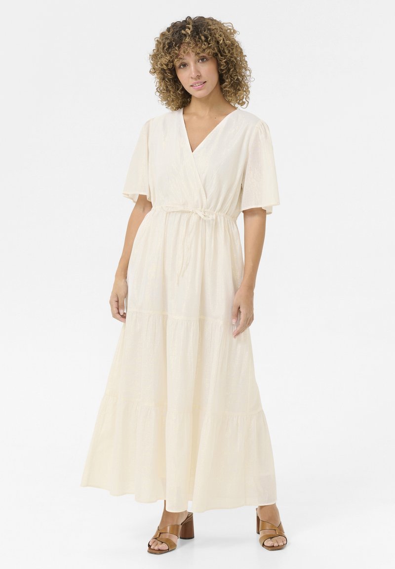 Light cream maxi dress with a V-neck, short flutter sleeves, and a tie waist. Features a tiered skirt with a soft, textured fabric. Brown heeled sandals.