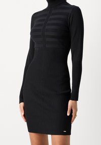 Robe noire côtelée à manches longues, avec un col montant et des détails à rayures horizontales sur le corsage, silhouette ajustée.