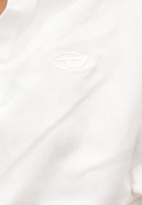 Λευκό ύφασμα με λεπτή ανάγλυφη κεντητική σε σχήμα οβάλ κοντά στο κέντρο, που δείχνει την υφή και τις ραφές στο ύφασμα.