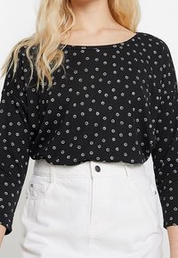 Schwarzes Langarmoberteil mit weißen kreisförmigen Mustern und weitem Ausschnitt. Der Stoff wirkt weich und wird mit hoch taillierten weißen Hosen kombiniert.