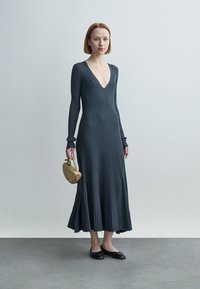 Σκούρο γκρι ριμπ πλεκτό φόρεμα με μακριά μανίκια και βαθύ V-λαιμόκοψη, φουσκωτή φούστα, σετ με μικρή μπεζ τσάντα χειρός και μαύρες μπαλαρίνες.