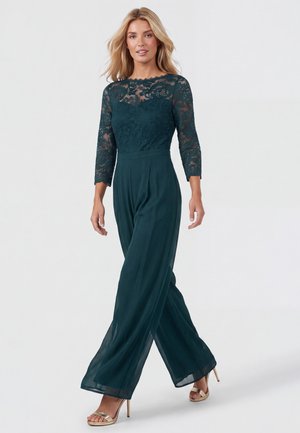 Femme marchant dans une combinaison en dentelle vert foncé avec des manches trois-quarts et un pantalon large, portant des sandales dorées à talons hauts.