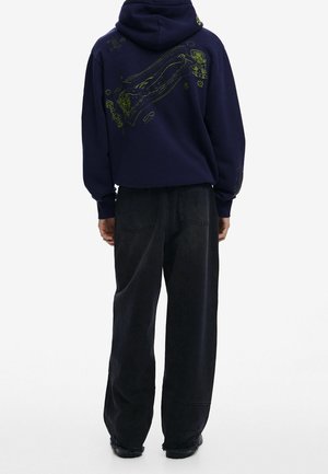 Marineblauwe hoodie met groen grafisch ontwerp op de rug, in een relaxed fit. Gecombineerd met losse zwarte denim broek en zwarte schoenen.