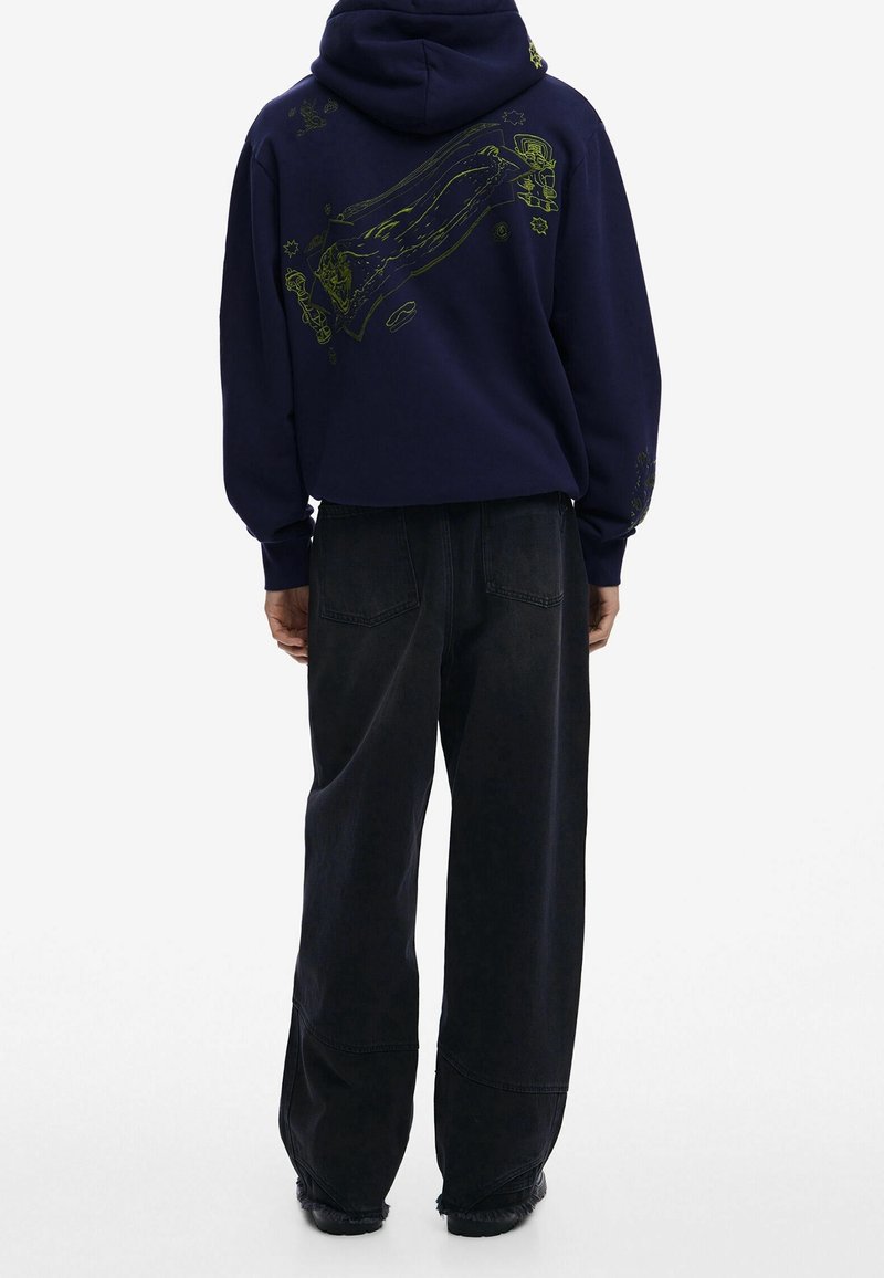 Granatowy sweatshirt z zielonym grafikiem na plecach, w luźnym kroju. W połączeniu z luźnymi czarnymi dżinsami i czarnymi butami.