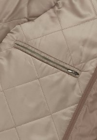 Beżowa tkanina pikowana o gładkiej fakturze; posiada zamykaną na zamek kieszeń z szarym futerkiem w środku i kontrastowym brązowym wykończeniem.