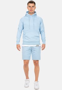 Haut à capuche bleu clair avec cordons de serrage et poche avant, associé à un short assorti. Le tissu est doux et la coupe est décontractée. Des baskets blanches complètent le look.