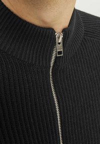 Schwarzer gerippter Pullover mit hohem Kragen, der über ein silbernes Reißverschlussdetail verfügt und aus strukturiertem Stoff für Wärme und Komfort gefertigt ist.