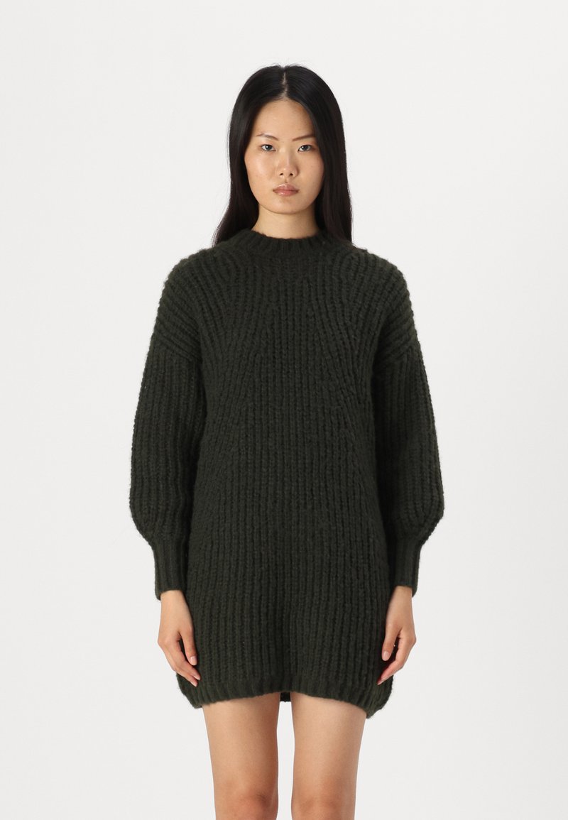 Abito maglione lavorato a maglia spessa di colore verde scuro. Presenta una texture a coste, collo alto e maniche a palloncino. Leggermente più lungo sul retro.