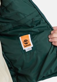 Grön nylonjackas insida med en vit och orange Timberland-logotyp på lappen, med ett blixtlåsfack och slät textur.