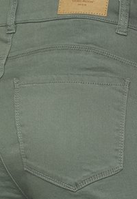 Vaqueros de denim verde con un acabado texturizado, que cuentan con un bolsillo trasero y una etiqueta de cuero marrón con la marca "VERO MODA" en relieve.