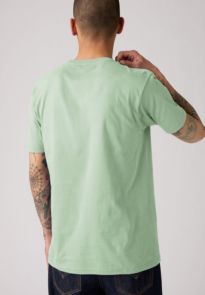 Mann mit Tätowierungen, der ein hellgrünes T-Shirt und dunkle Jeans trägt, von hinten gezeigt, richtet mit der rechten Hand den Ausschnitt vor einem einfarbigen Hintergrund.