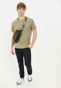 Lys olivengrøn t-shirt, tætsiddende, suppleret med mørkeblå bukser. Sort crossbody taske med lynlåsdetalje. Modellen bærer hvide sneakers.