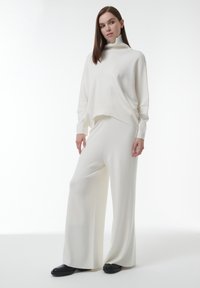 Witte gebreide turtleneck trui met een losse fit, gecombineerd met witte wijde broekspijpen. De stof lijkt zacht en textuur te hebben. Zwarte schoenen maken de outfit compleet.