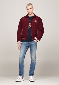 Giacca bomber in suede bordeaux con strisce bianche sui polsini e l'orlo, logo ricamato sul petto, abbinata a jeans blu a gamba affusolata e sneakers bianche.