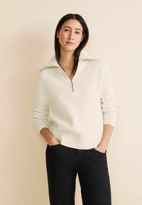 Pull en maille blanc avec un col haut et une fermeture �éclair à l'avant. Manches longues et ourlet arrondi, associé à un pantalon sombre. Texture lisse et côtelée.