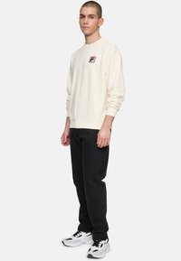 Sudadera de color crema con puños y dobladillo acanalados, que presenta un pequeño logo bordado. Combinada con jeans negros y zapatillas blancas.