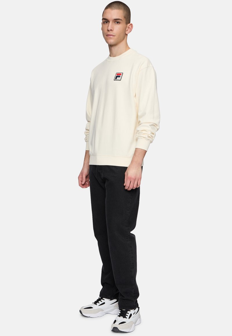 Felpa color crema con polsini e orlo a coste, caratterizzata da un piccolo logo ricamato. Abb binata a jeans neri e sneakers bianche.