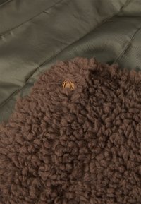 Brun, textured fleece stof overlapper glat, mørkegrøn nylon, med en rund brun knap fastgjort til fleece.