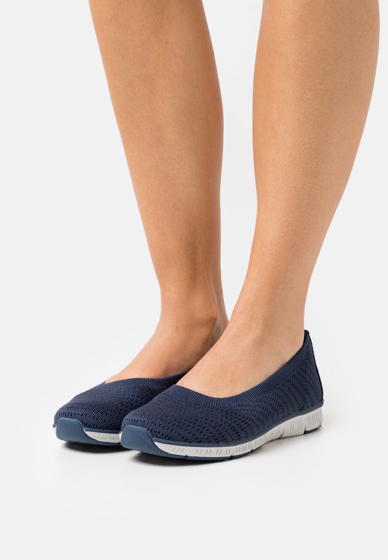 Pieds portant des chaussures slip-on en maille bleu marine avec semelles blanches flexibles sur fond blanc.