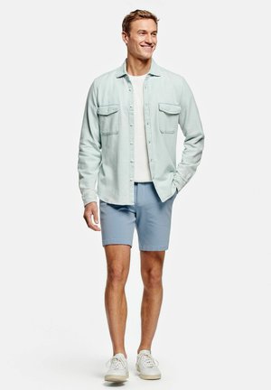 Jeune homme souriant, portant une chemise bleu clair à boutons sur un t-shirt blanc, un short bleu et des baskets blanches, debout sur un fond blanc.