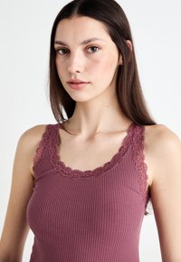Top a canotta a coste di un profondo colore malva, con finiture in pizzo lungo lo scollo e le spalline, dalla silhouette aderente e morbida al tatto.