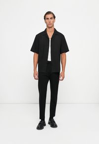 Black short-sleeve button-up shirt paired with black drawstring trousers and black slip-on shoes. Simple, solid design with a relaxed fit.