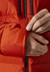 Červená zateplená bunda s texturovaným povrchem. Ruka upravuje uzávěr na suchý zip na manžetě, což zdůrazňuje bezpečné uchycení a detail designu.