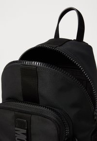 Čierny látkový batoh s dvoma zipsovými priehradkami, z ktorých jedna je čiastočne otvorená, a s horným uchom na nosenie na jednoduchom svetlom pozadí.