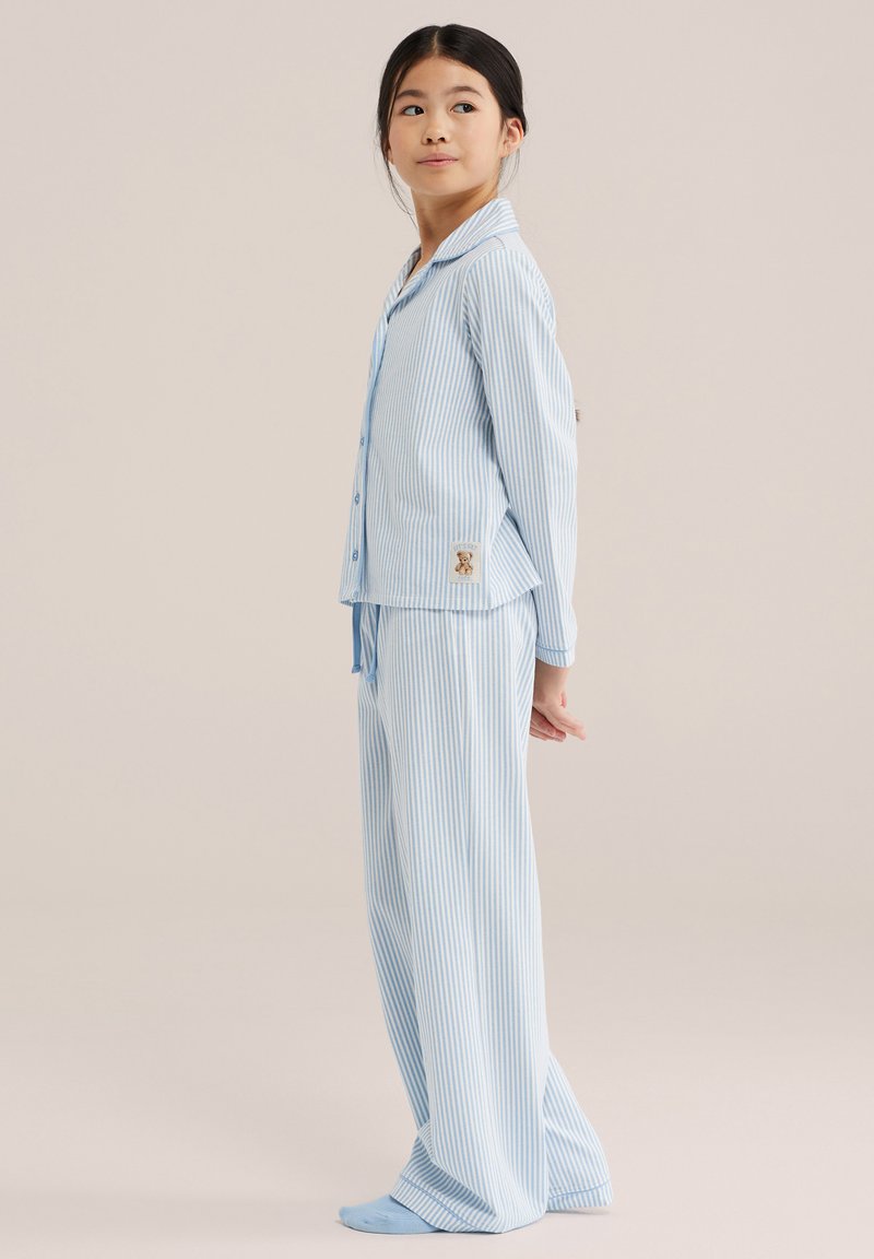 Jeune fille portant un pyjama rayé bleu et blanc à manches longues et un pantalon assorti, debout avec les mains derrière le dos, regardant sur le côté.