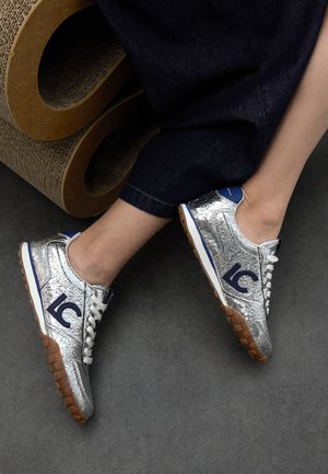Zapatillas plateadas con un acabado metálico agrietado, cordones blancos, acentos azules y una suela de goma texturizada. Presentan un logo azul en el lateral.