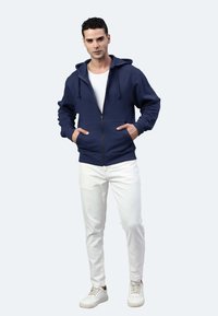 Felpa con zip color blu navy realizzata in morbido tessuto, presenta un cappuccio, lacci e tasche frontali, abbinata a jeans bianchi e sneakers.