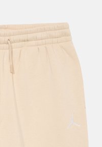 Pantaloni de trening bej cu talie elastică și șnur ajustabil, având un mic logo Air Jordan Jumpman alb pe partea stângă.