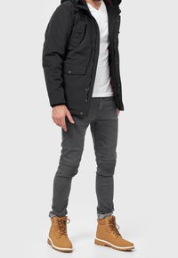 Svart vadderad jacka med röd foder, grå slim fit-jeans med ribbade detaljer och beige snörkängor med vita sulor.