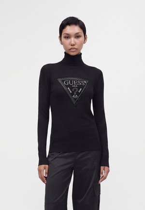 Femme aux cheveux courts portant un pull à col roulé noir avec un grand logo triangulaire Guess et un pantalon noir sur un fond uni.