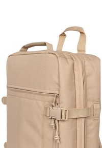 Tan rektangulär ryggsäck gjord av texturerad tyg, med ett topphandtag, framficka med dragkedja och säkerhetsrem med hårdvarufästen.