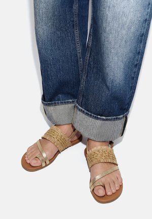 Pies con sandalias de tiras tejidas y metálicas en tono marrón claro, combinadas con vaqueros azules remangados sobre un fondo blanco.