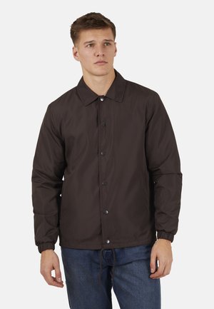 Młody mężczyzna ubrany w brązową, zapinaną na guziki kurtkę z kołnierzem i elastycznymi mankietami, w połączeniu z niebieskimi dżinsami, stojący na białym tle.