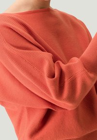 T-shirt à manches longues côtelé en tissu corail, avec un col large et une texture douce, des manches amples et drapées créant une silhouette décontractée.
