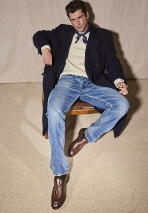 Manteau marine croisé, pull en tricot crème, jean en denim bleu et mocassins en cuir marron avec des franges. Assis sur une chaise en bois.