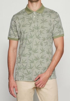 Mand iført lysegrøn kortærmet poloshirt med tonalt bladmønster og beige bukser, stående mod en ensfarvet baggrund.